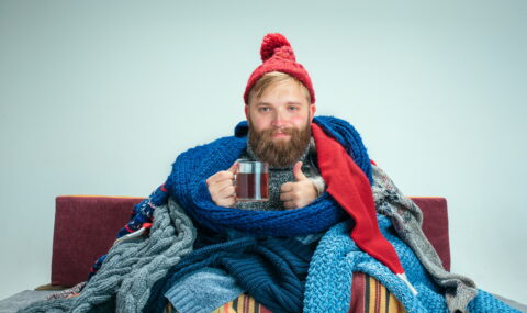 bearded-sick-man-with-flue-sitting-sofa-home-studio-with-cup-tea-covered-with-knitted-warm-clothes-illness-influenza-concept-relaxation-home-healthcare-concepts